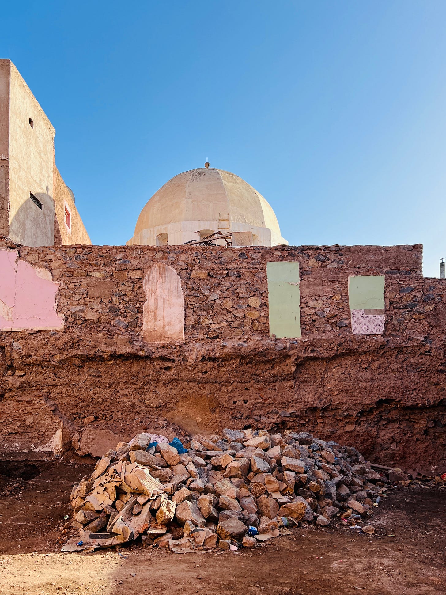 Partially demolished wall in Marrakech with painted traces of former rooms and window shapes, rubble piled in front, and a dome rising behind against a clear blue sky. Partially demolished wall in Marrakech with painted traces of former rooms and window shapes, rubble piled in front, and a dome rising behind against a clear blue sky.