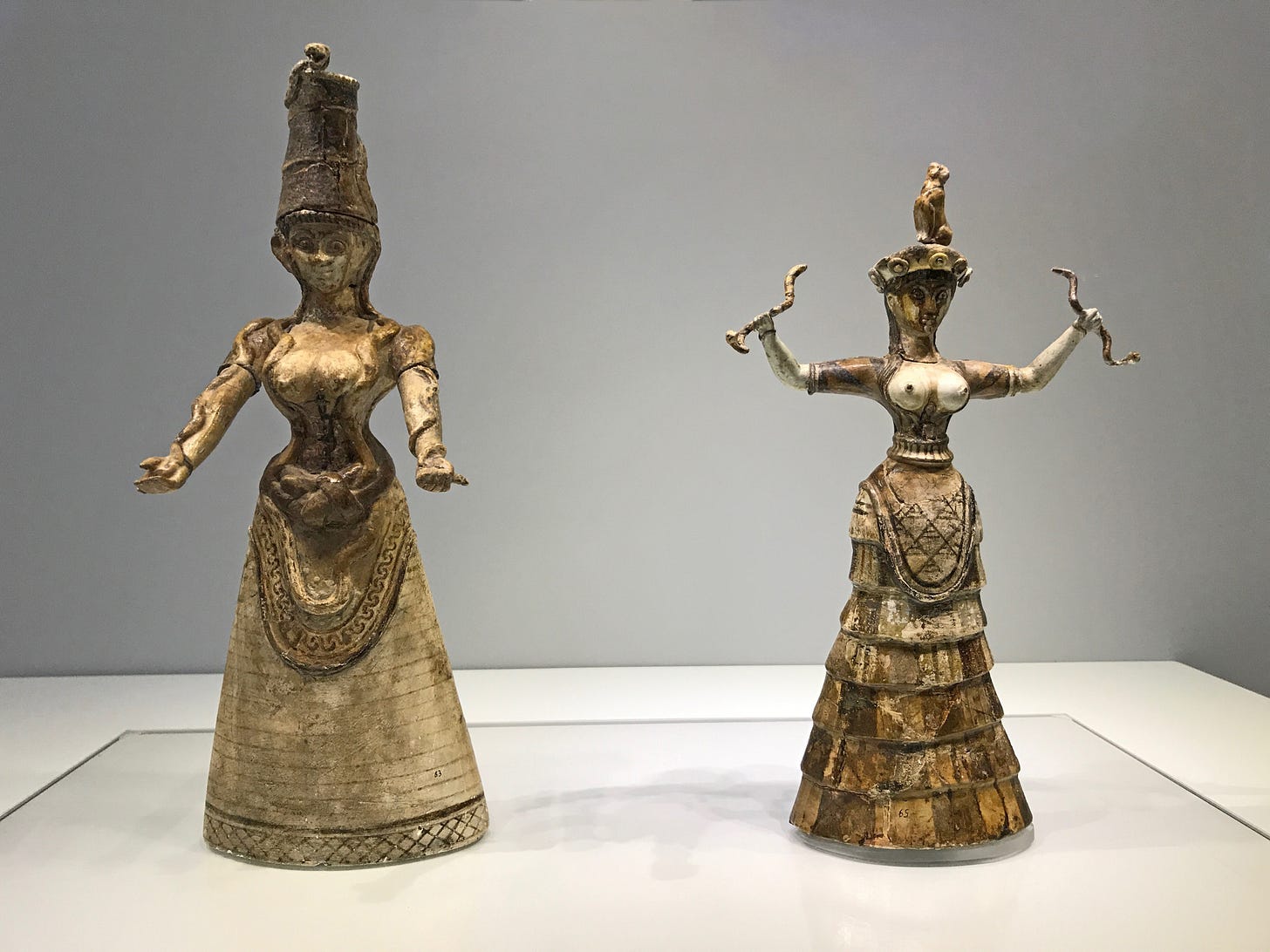 Two Minoan faience figurines of Snake Goddesses, both wearing long skirts and short-sleeved tops that leave their breasts bare. The one on the left is slightly taller than the one on the right. The one on the left has a tall cylindrical hat with a snake wrapped around it as well as snakes twining up her arms and around her waist. The one on the right has a beret-style hat with a cat sitting on it. She holds a writhing snake in each outstretched hand.