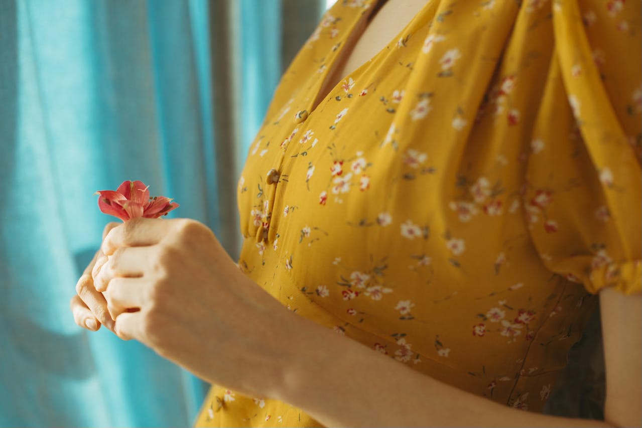 Cropped side view of woman in mustard dress holding a red flower in front of her chest. Cropped side view of woman in mustard dress holding a red flower in front of her chest.
