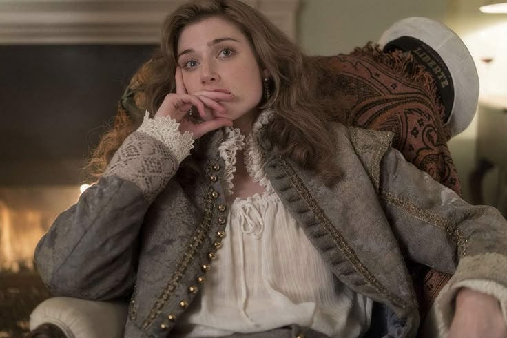 Elizabeth Debicki como Virginia Wolf. ela está sentada na cadeira com uma mão apoiando o queijo e o dedo cobrindo a boca. sua expressão é reflexiva