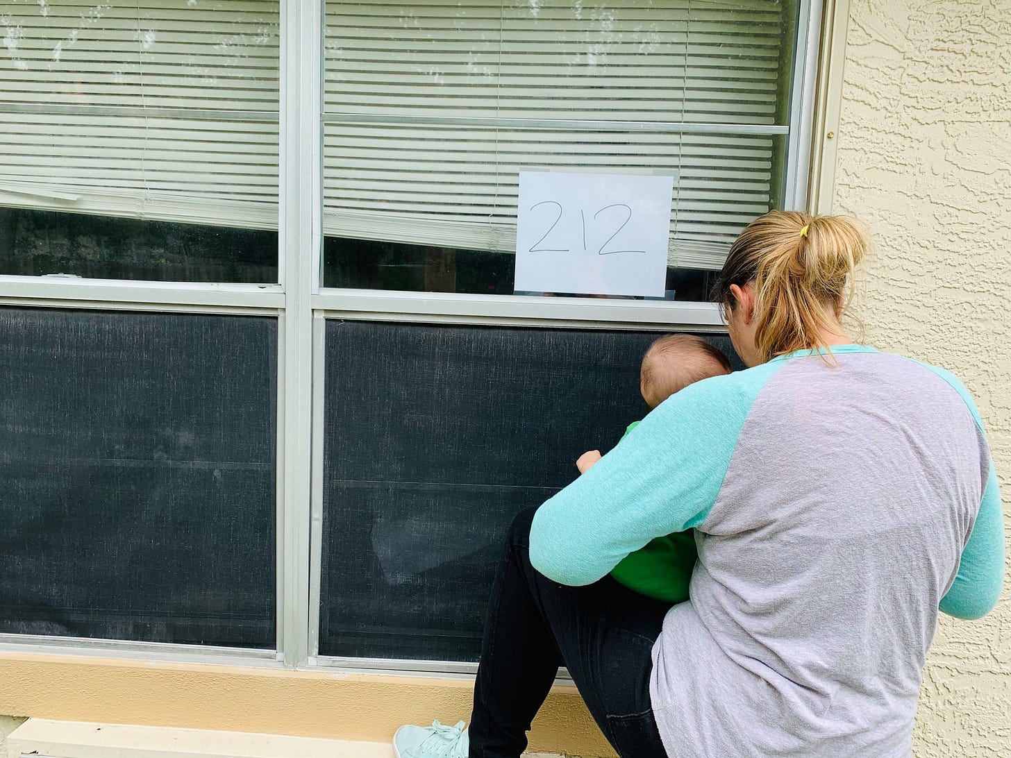 A mom holds a baby up to a darkened window. The blinds are half down, and there's a handwritten room number stuck to the glass. A mom holds a baby up to a darkened window. The blinds are half down, and there's a handwritten room number stuck to the glass.