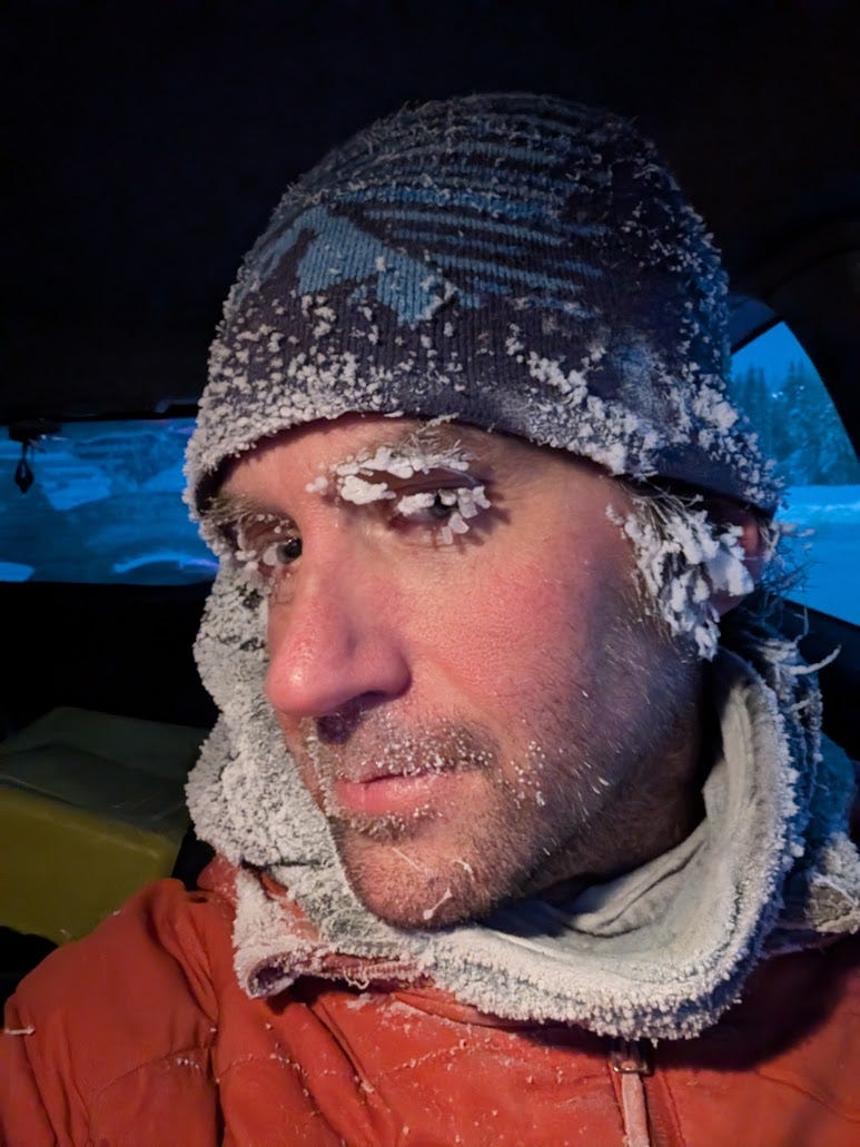 A selfie taken inside a car at night. A man wearing a blue knit hat and orange jacket has heavy frost coating his eyelashes, eyebrows, beard stubble, and fleece neck gaiter. His cheeks are flushed red from the cold. Snow-covered trees are faintly visible through the car window behind him. Fairbanks, Alaska.