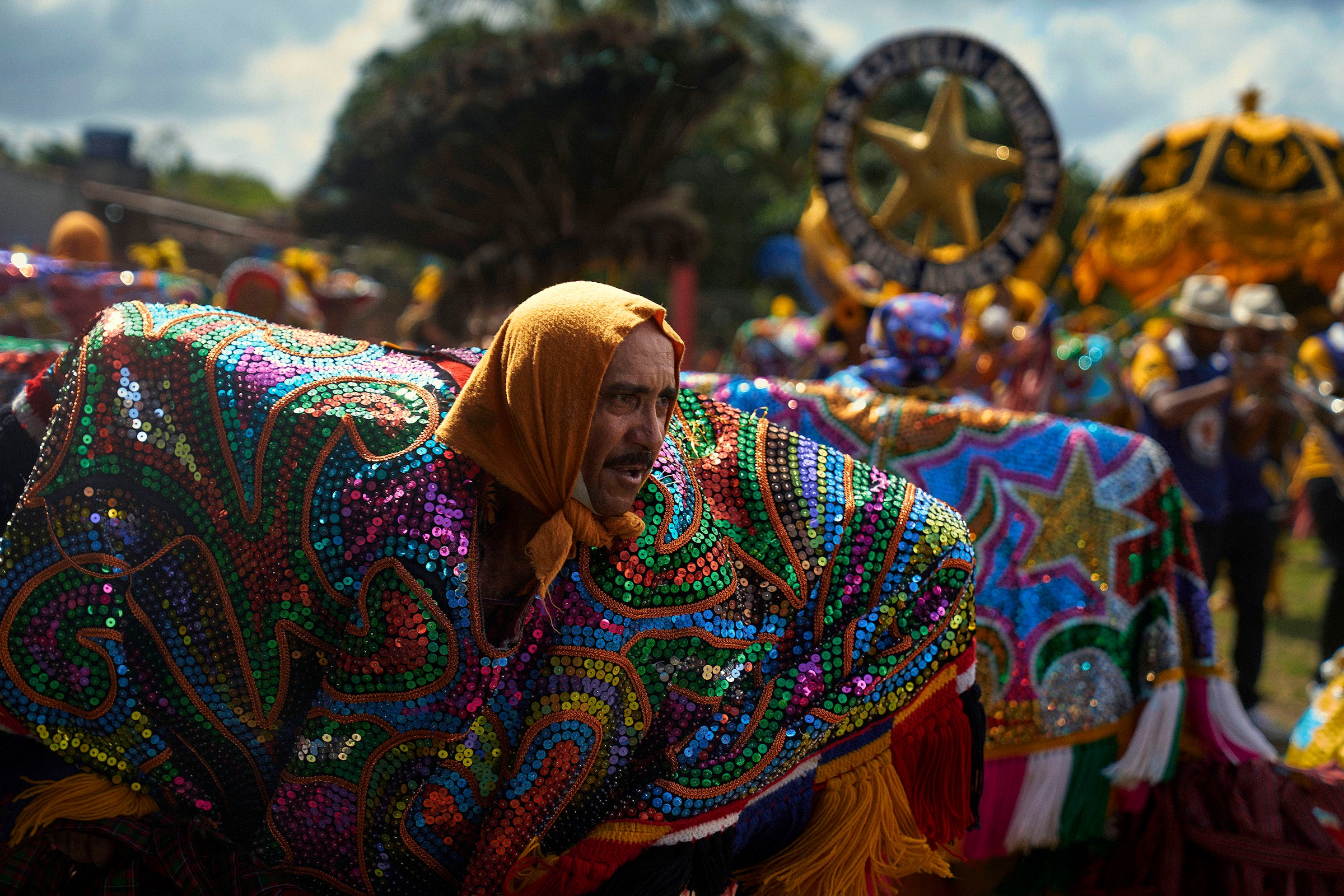A person in a colorful, sequined costume with a yellow headscarf participates in a festive outdoor parade, surrounded by others in vibrant attire and decorated floats under a bright sky.