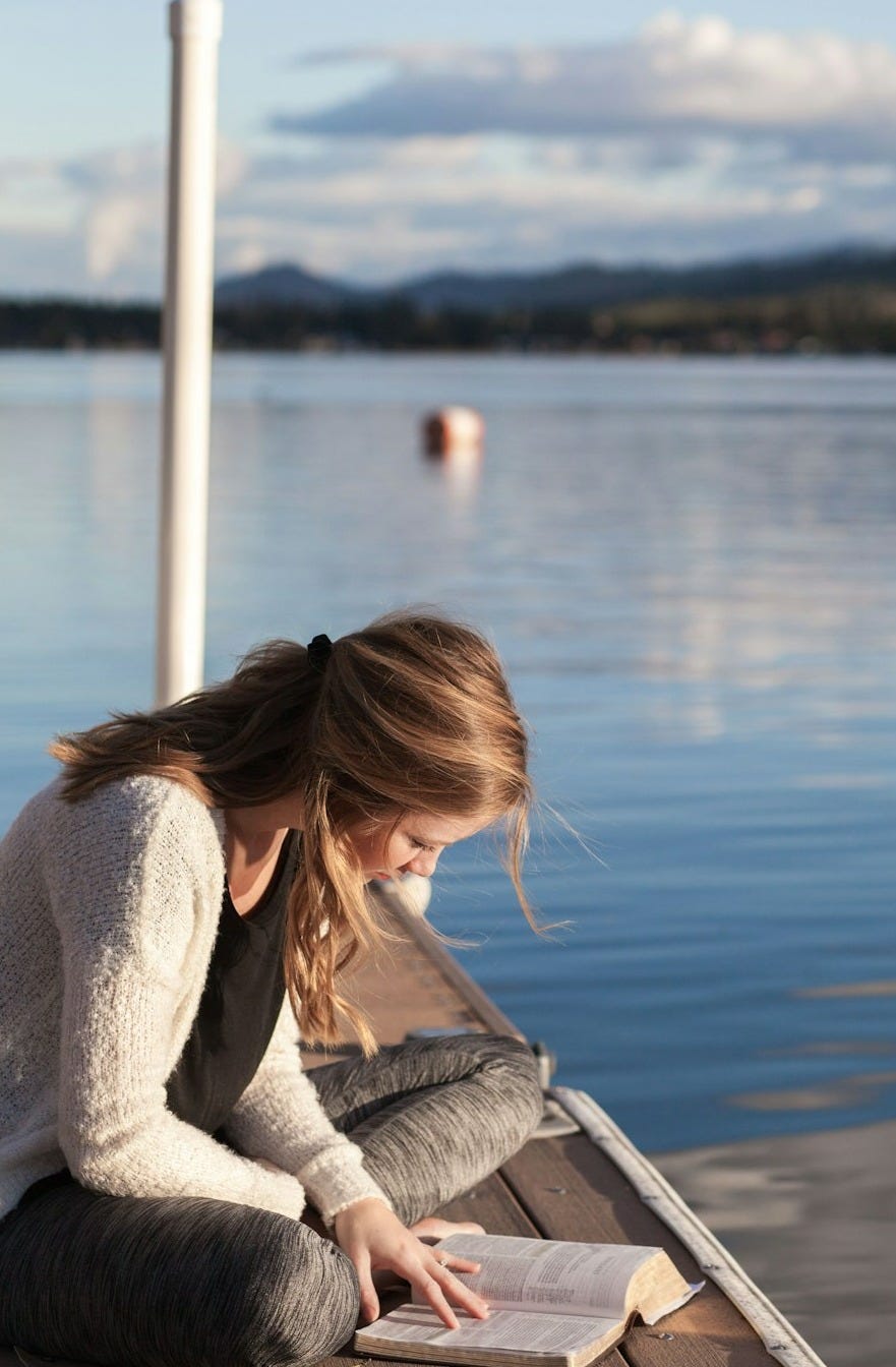 photo of woman reading book near body of water