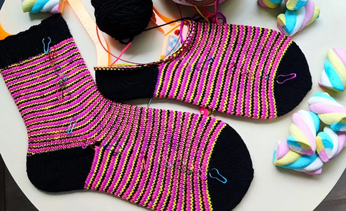 two striped socks on a table. the stripes are pink, yellow and red. one of the socks hasn't been completely knitted.