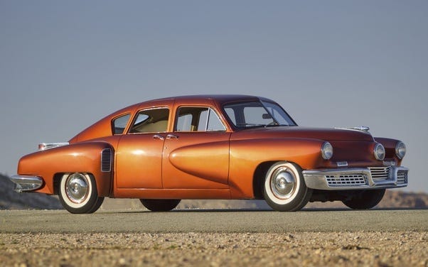 La imagen muestra un coche antiguo, de mediados del siglo XX, con un diseño elegante y una brillante pintura naranja. El vehículo está posicionado en lo que parece ser una carretera abierta o un área despejada. El diseño clásico del coche, como las formas redondeadas de la carrocería, los detalles cromados y los neumáticos con banda blanca, lo hacen interesante. La imagen muestra un coche antiguo, de mediados del siglo XX, con un diseño elegante y una brillante pintura naranja. El vehículo está posicionado en lo que parece ser una carretera abierta o un área despejada. El diseño clásico del coche, como las formas redondeadas de la carrocería, los detalles cromados y los neumáticos con banda blanca, lo hacen interesante.