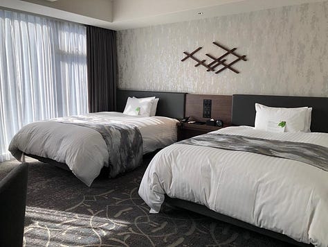A modern and spacious hotel room with twin beds, a seating area with armchairs and a chaise lounge, a flat-screen TV, and large windows with sheer curtains. The accessible bathroom features grab bars, a handheld showerhead, and a shower chair.
