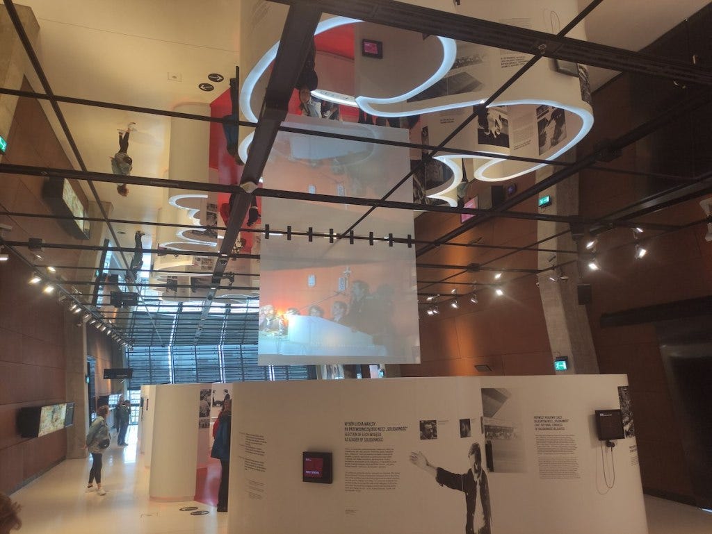 An exhibit stands in a room with a mirror-roof. That mirror allows you to see that the curved panels of the exhibition are that shape so they spell out the word "solidarnosc"