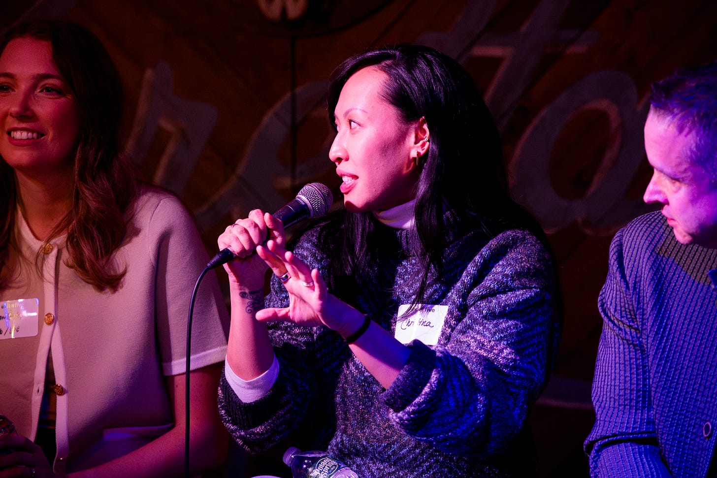 A woman speaking into a microphone while seated on a panel during a networking event. She is mid-sentence, gesturing with her hands, with two other panelists seated beside her. The lighting is warm and colorful, creating a lively atmosphere.