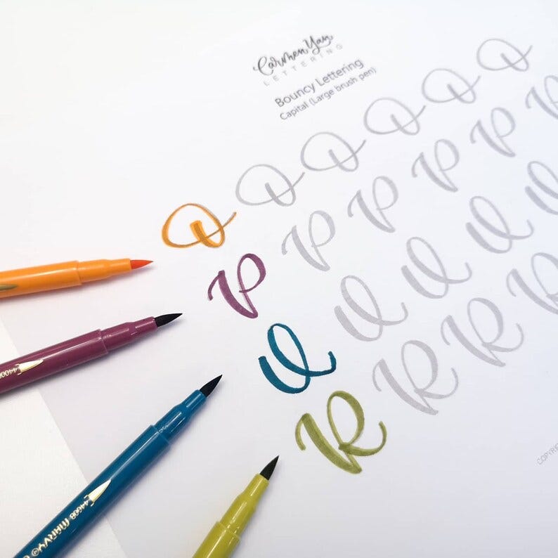May include: A sheet of paper with the words "Carmen Yay Lettering" and "Bouncy Lettering" at the top. The paper has several examples of the letter "P" written in different colors, including orange, purple, teal, and green. The text "Capital (Large brush pen)" is also visible.