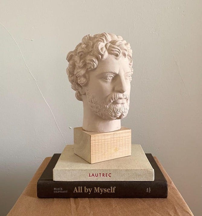 May include: A beige plaster bust of a man with curly hair sits atop a stack of books. The bust is on a light wood base. The top book is light beige with the word 'LAUTREC' printed on it. The bottom book is dark brown with the title 'All by Myself' printed in a serif font.