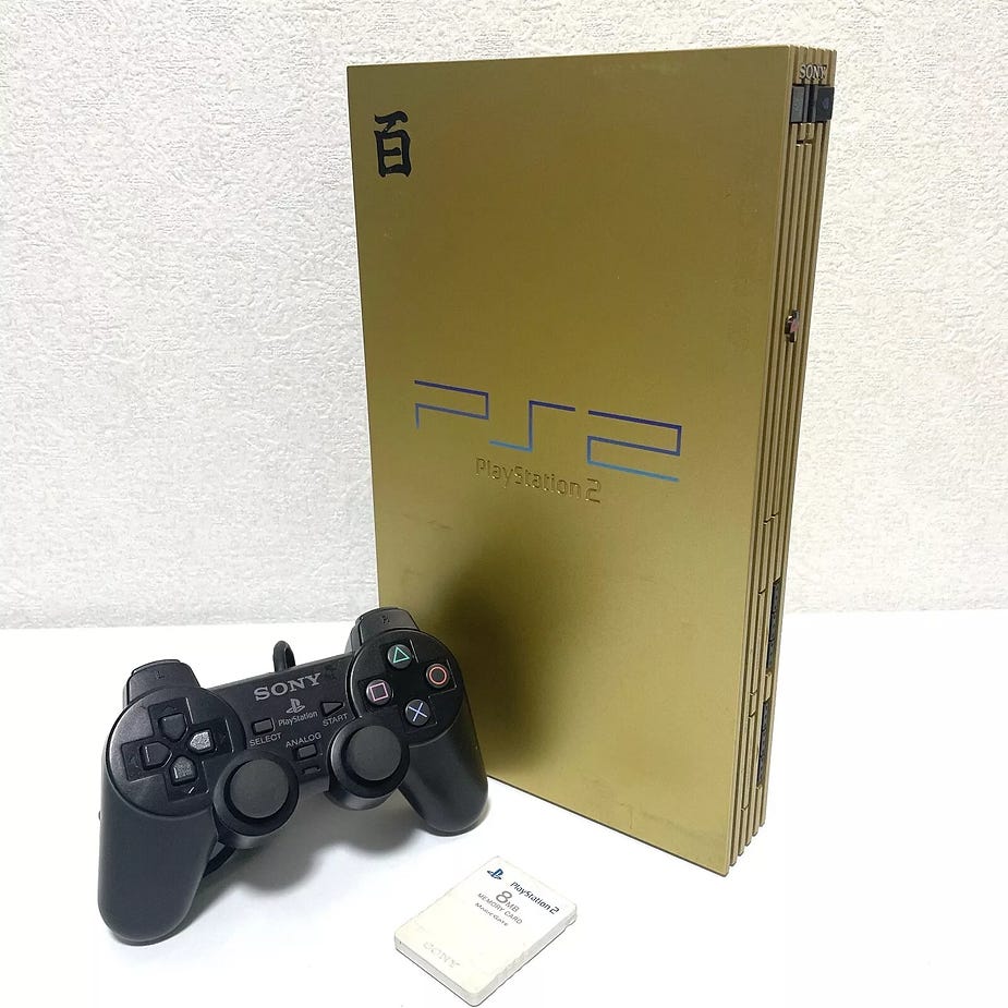 Gold PlayStation 2 with a black controller and memory card on a white surface. Blue "PS2" text on the console, Japanese character visible.