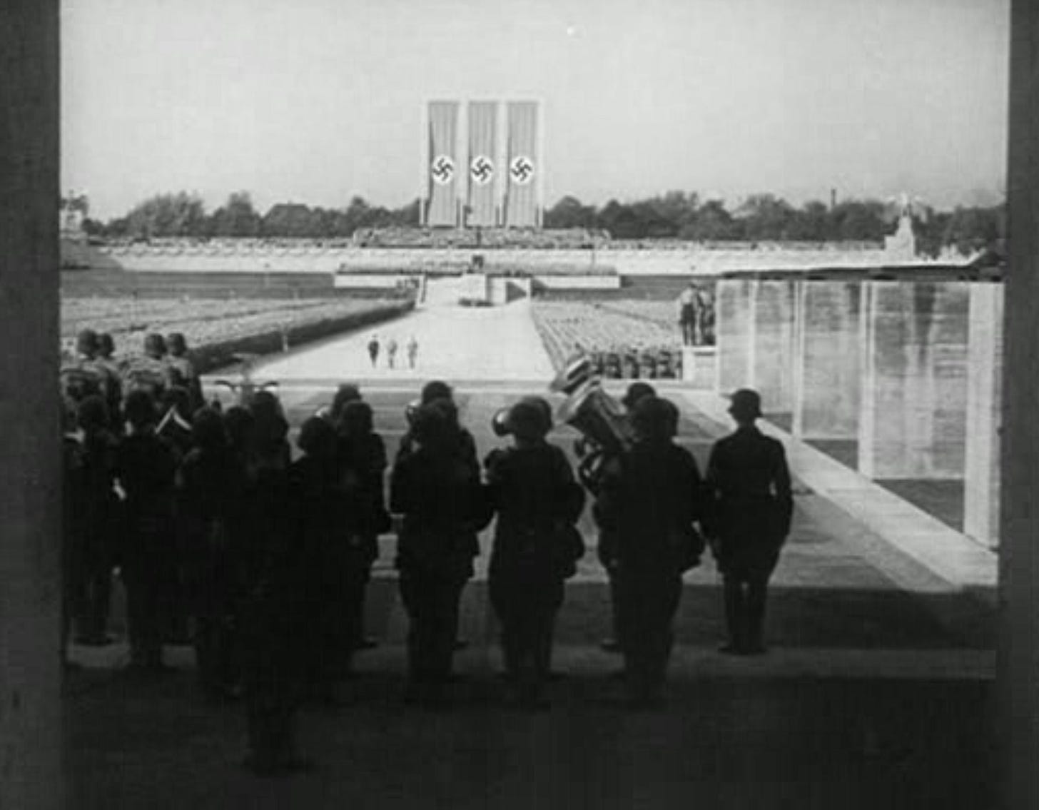 A frame from the film Triumph of the Will, looking across a huge rally ground from a raised platform. A brass band is in the foreground, backs to the camera. In the middle-ground, thousands of troops in neat rows stand at attention, with a wide path down the center.  Three small figures in a row walk down the pathway, toward the camera. In the background is an another raised speaking platform flanked by monumental steps. Three giant swastika banners hang over it, aligned with the pathway through the troops. There is a sense of the vast scale of the event.