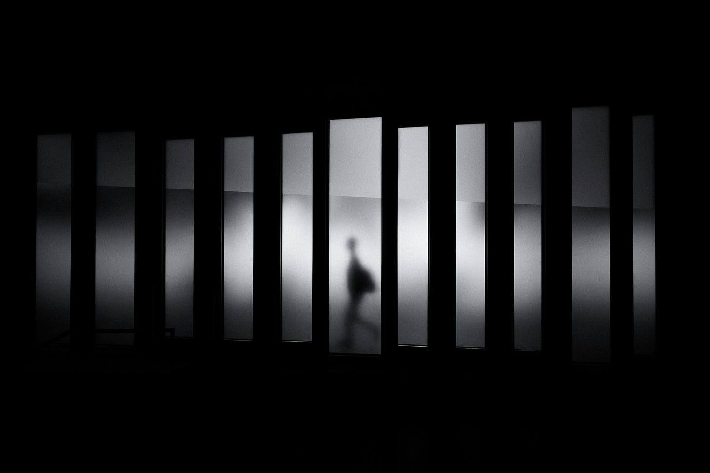 A high-contrast silhouette of a figure walking through a corridor of vertical light and shadow. A high-contrast silhouette of a figure walking through a corridor of vertical light and shadow.