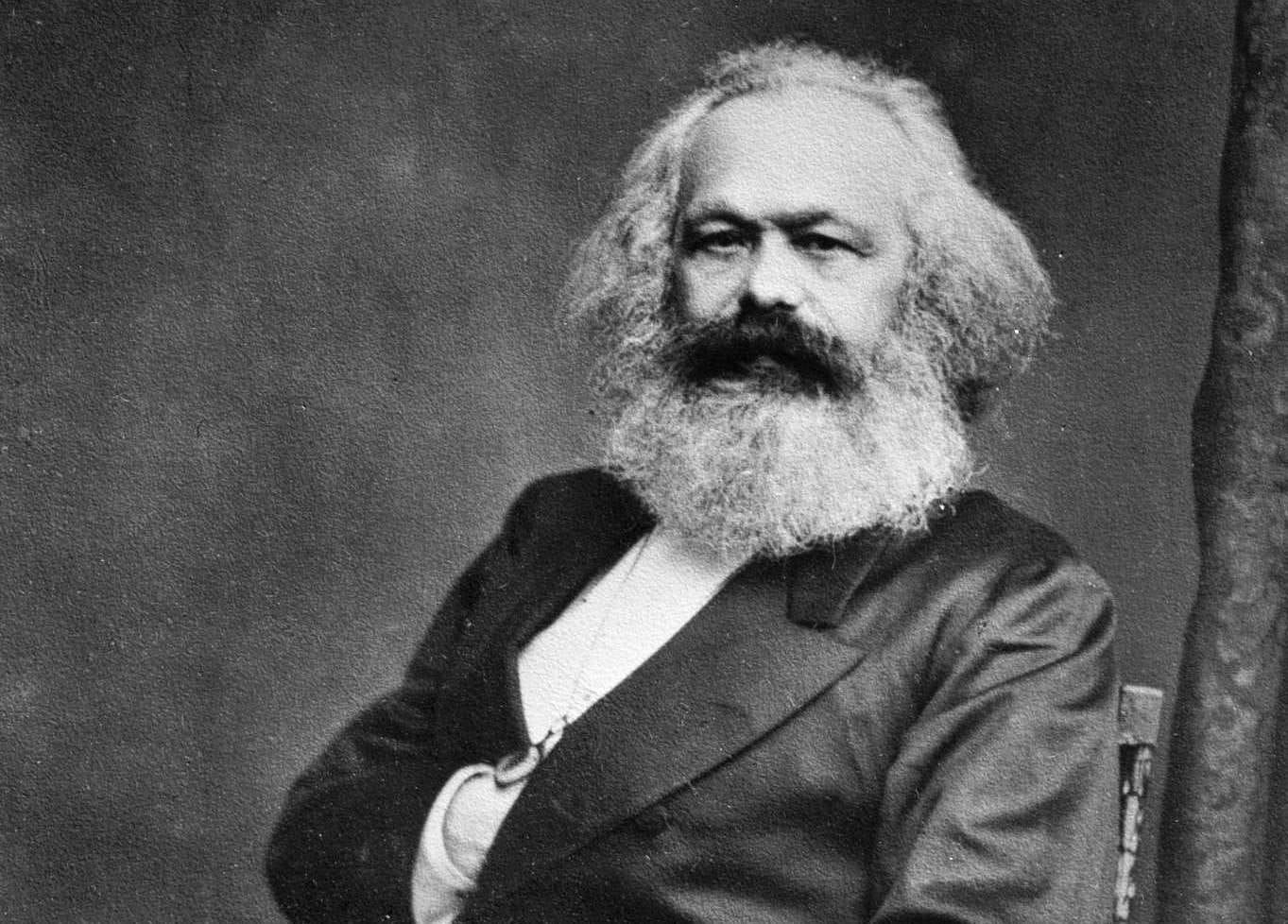 A bearded man with his hand tucked into his jacket sitting in a chair.