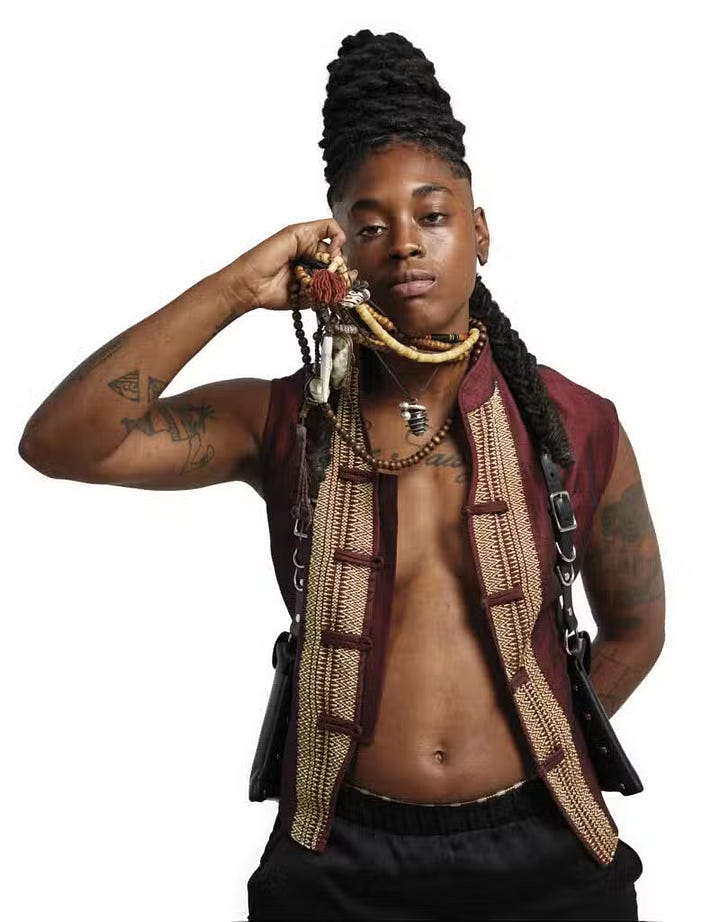 1) A person with a confident expression poses against a white background. They wear a maroon vest with an intricate trim, black pants, and they have styled hair. Tattoos adorn their arms, and they hold a beaded necklace near their face. Their vest is open, revealing their bare chest, and they are staring straight into the camera. 2) A person with sunglasses and braided hair stands confidently on a rooftop, wearing a white leather jacket and gray sweater. The background shows a skyline under a clear sky. They are holding their hands together in front of their torso, and they are staring straight into the camera with a neutral expression.