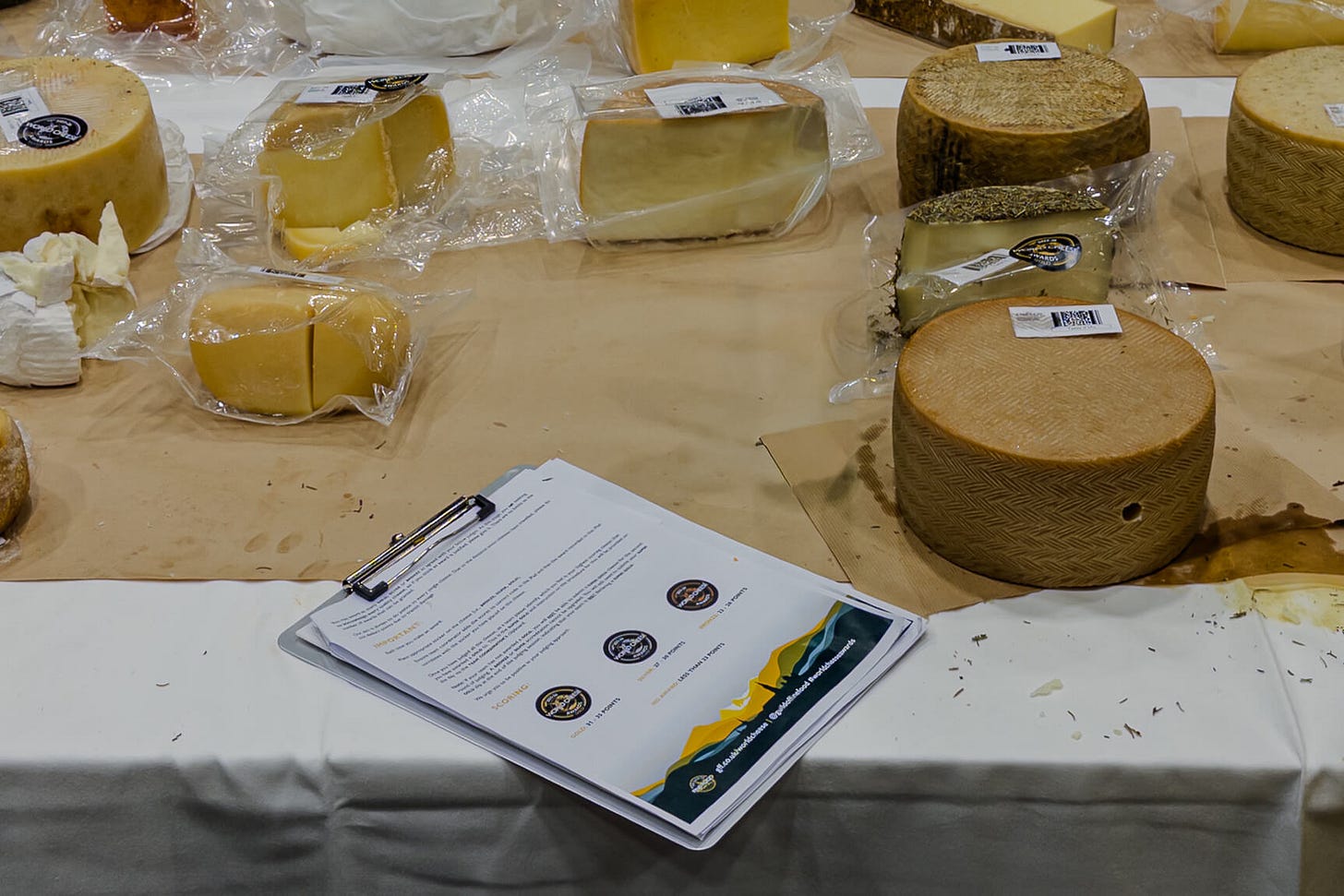 Ein Jury-Tisch bei den World Cheese Awards ist mit verschiedenen Käselaiben und angeschnittenen Proben bedeckt, viele davon vakuumverpackt und mit Etiketten versehen. Rechts liegt ein grosser runder Laib mit gemusterter Rinde, daneben kleinere Stücke und Spuren von Schnittresten. Links unten auf dem Tisch liegt ein Klemmbrett mit Bewertungsunterlagen der Jury.