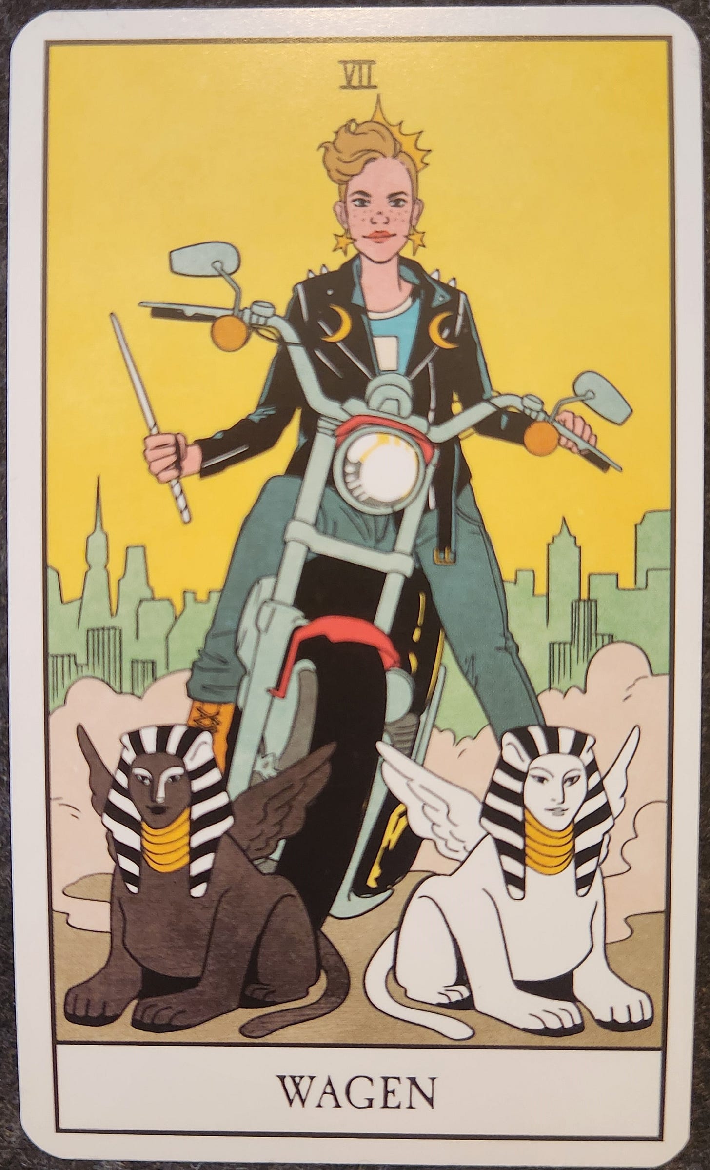 A tarot card labelled XII  Wagen. A person with short choppy hair is straddling a motorcycle. Though the motorcycle is not in motion, there is dust billowing behind it as though the engine is on. The person is wearing a black leather jacket, adorned with two crescent moons. They are wearing a golden crown and golden star earrings. They are holding a wand in one hand. In front of the motorcycle are two sphinxes, one black and one white. In the distant background, is a cityscape. 