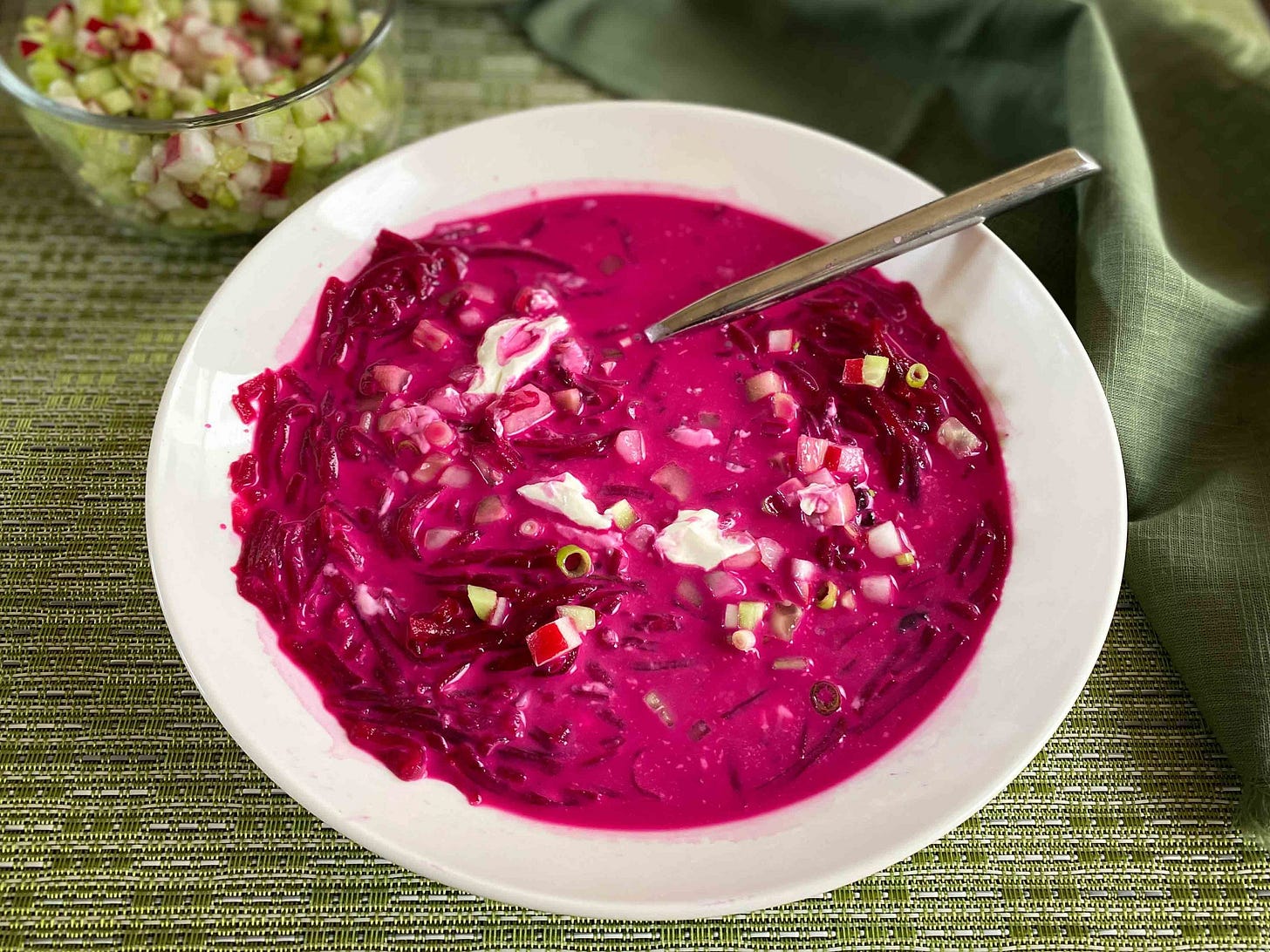 A bowl of cold beet borscht with sour cream mixed in, garnished with diced cucumber radishes and scallions