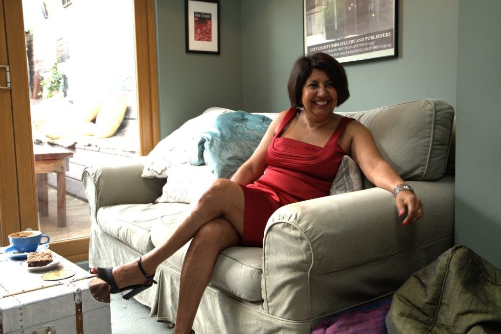 A woman of Indian heritage with a black bob is wearing a chic red satin dress and black heels. She leans back on a pale green sofa – away from a coffee table with a blue mug of coffee.
