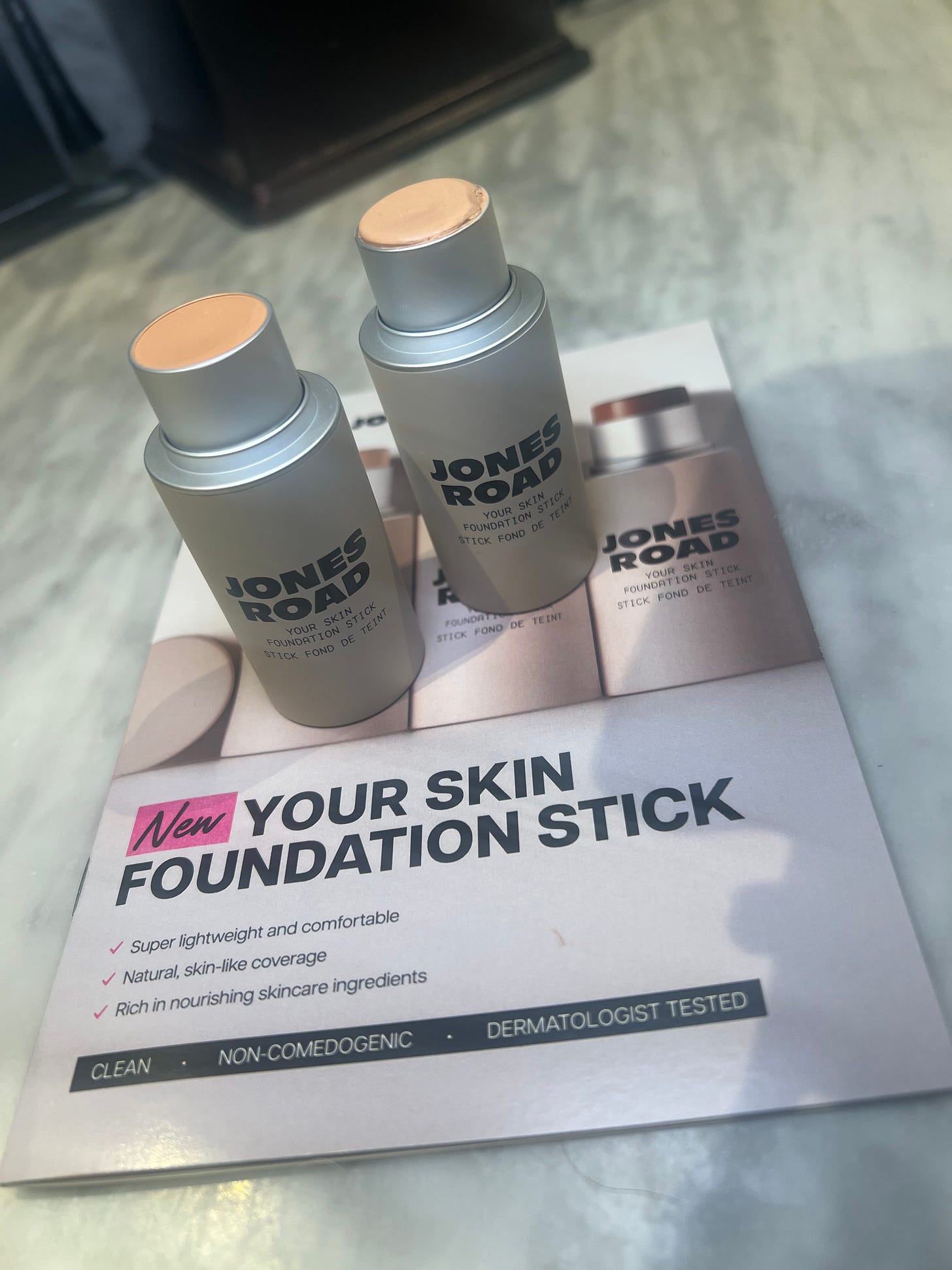 Two silver Jones Road "Your Skin" Foundation Sticks stand upright on top of a promotional product card. The sticks are uncapped, revealing creamy, skin-toned product. The background is a grey marble surface, highlighting the sleek, minimalist packaging of the brand.