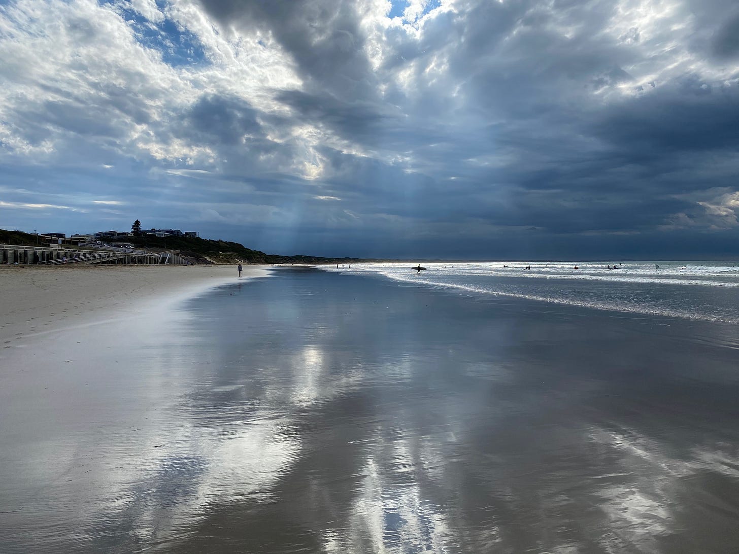 Beach scene, early morning. A wide-angle shot of Ocean Grove beach, with beams of sunlight from a mostly cloudy sky. On the right, the surf, on the left, the dunes. The light is reflected strongly in the wet sand. A few swimmers and a surfer with board under arm are in the middle distance.