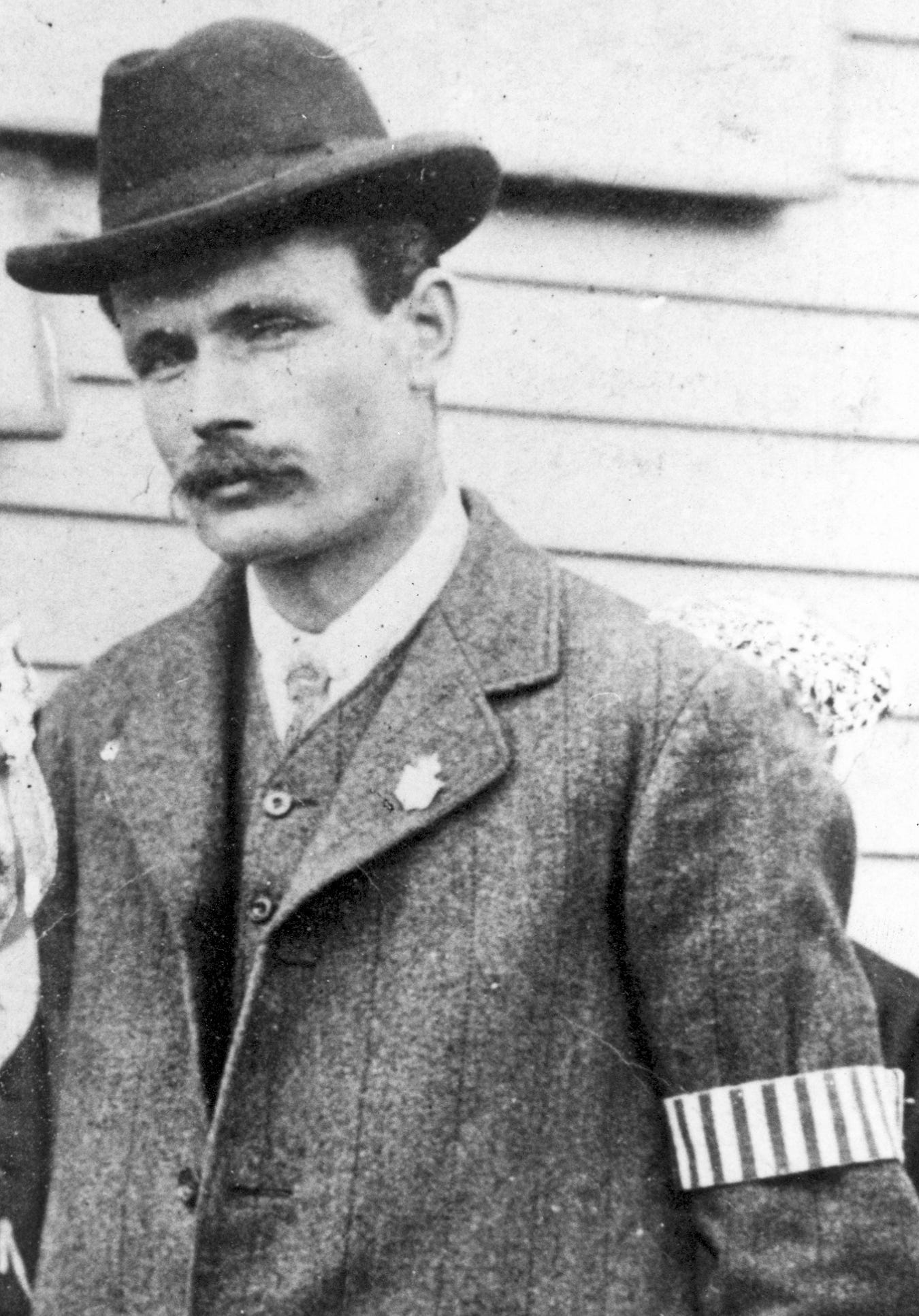 Black and white photo of a man in a shirt, tie, waistcoat and jacket wearing a fedora hat. He is of slim build and has a moustache. His striped Special Constable armband can be seen on his left arm.
