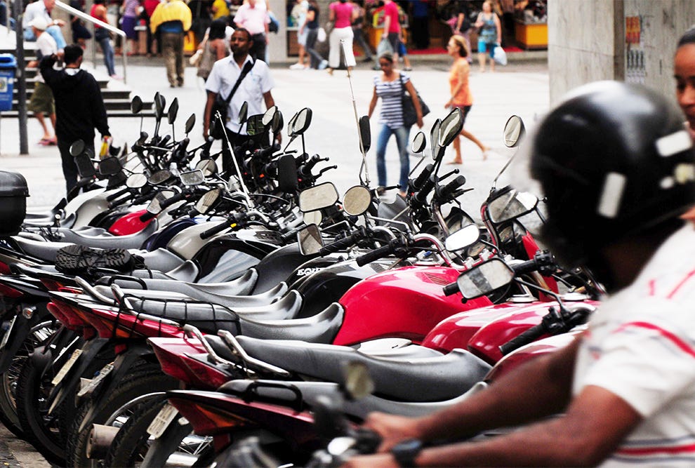 Fileira de motocicletas vermelhas e pretas estacionadas em primeiro plano com pessoas sentadas em algumas delas, enquanto ao fundo uma multidão circula em uma praça pública com edifícios, estruturas metálicas e pessoas em pé observando a cena. Fileira de motocicletas vermelhas e pretas estacionadas em primeiro plano com pessoas sentadas em algumas delas, enquanto ao fundo uma multidão circula em uma praça pública com edifícios, estruturas metálicas e pessoas em pé observando a cena.