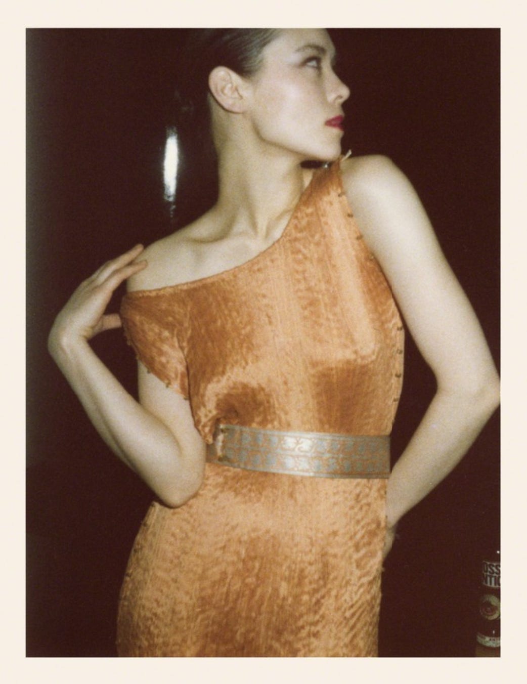 A short-haired woman with fair skin looks to one side and dramatically poses her arms while wearing a pale peach-colored silk dress with a contrasting stenciled velvet belt.