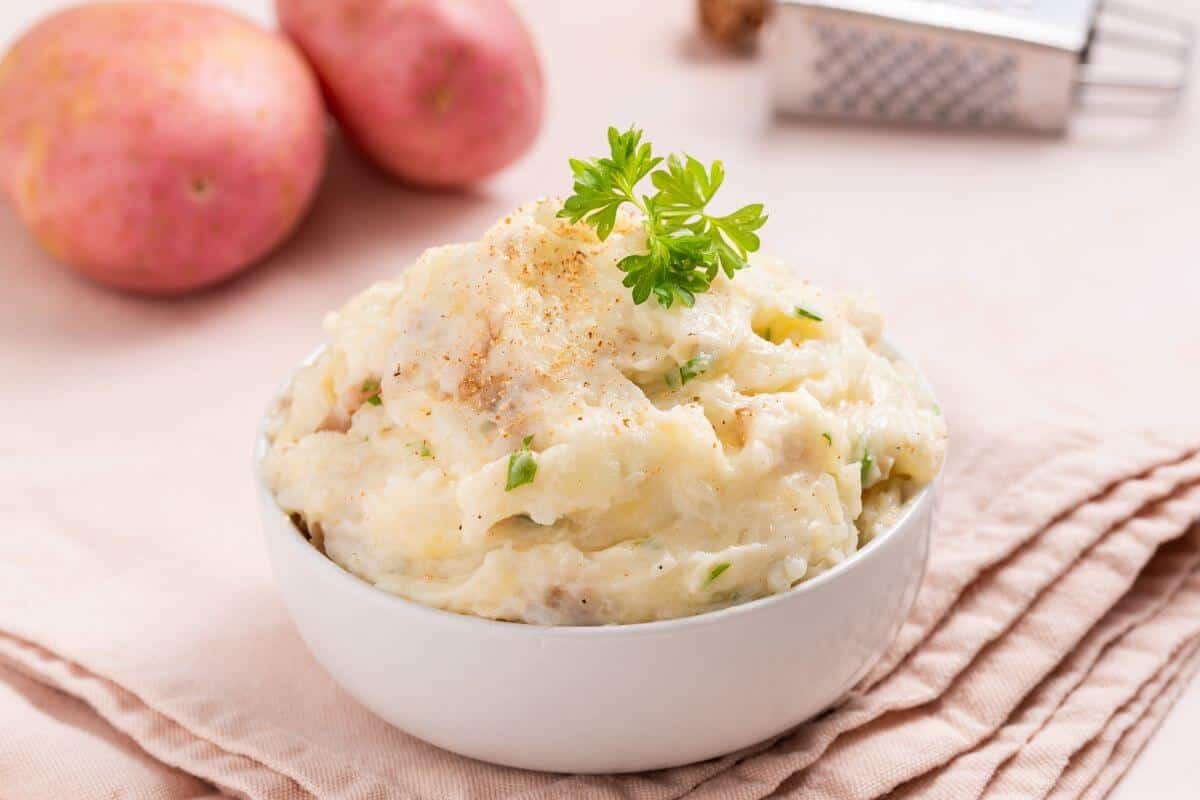 A bowl of mashed potatoes garnished with parsley, placed on a light-colored cloth next to a couple of whole potatoes. A grater is visible in the background.