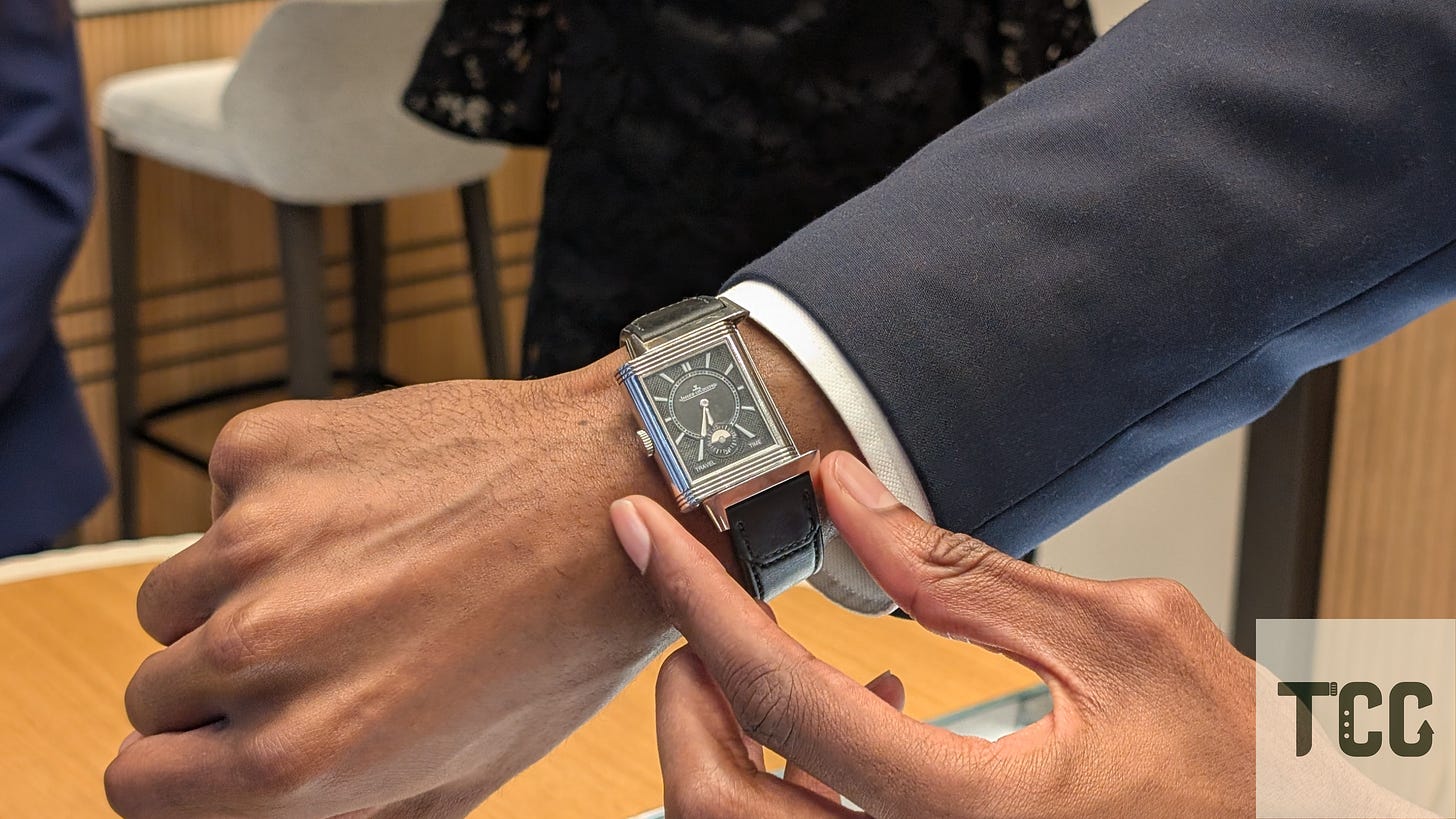 A wrist shot of a Jaeger-LeCoultre Reverso on a black leather strap. The wearer is dressed in a navy suit, pointing to the rectangular watch with polished case and black dial, standing at a boutique counter.