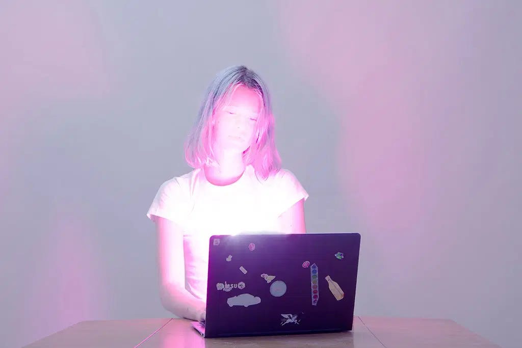 Uma menina branca de cabelos muito loiro descolorido na altura do ombro olha pra tela de seu laptop. Sentada numa cadeira que não vemos, com o laptop numa mesa branca, seu olhar é lânguido. Vemos as costas do laptop, escuro com vários adesivos colados. Ele é a única coisa escura na foto. Da sua tela, sai uma luz branca e rosa que colore tudo na imagem. A menina parece um ser de luz, toda branca,  com seus cabelos e delineado do corpo rosa. Seu rosto é quase todo branco. Se vê os olhos pequenos olhando pra tela e muito suavemente um traço de boca ou nariz, mas sua feição está quase toda tomada pela luz da tela. O fundo é vazio, iluminado apenas entre o rosa e o lilás.