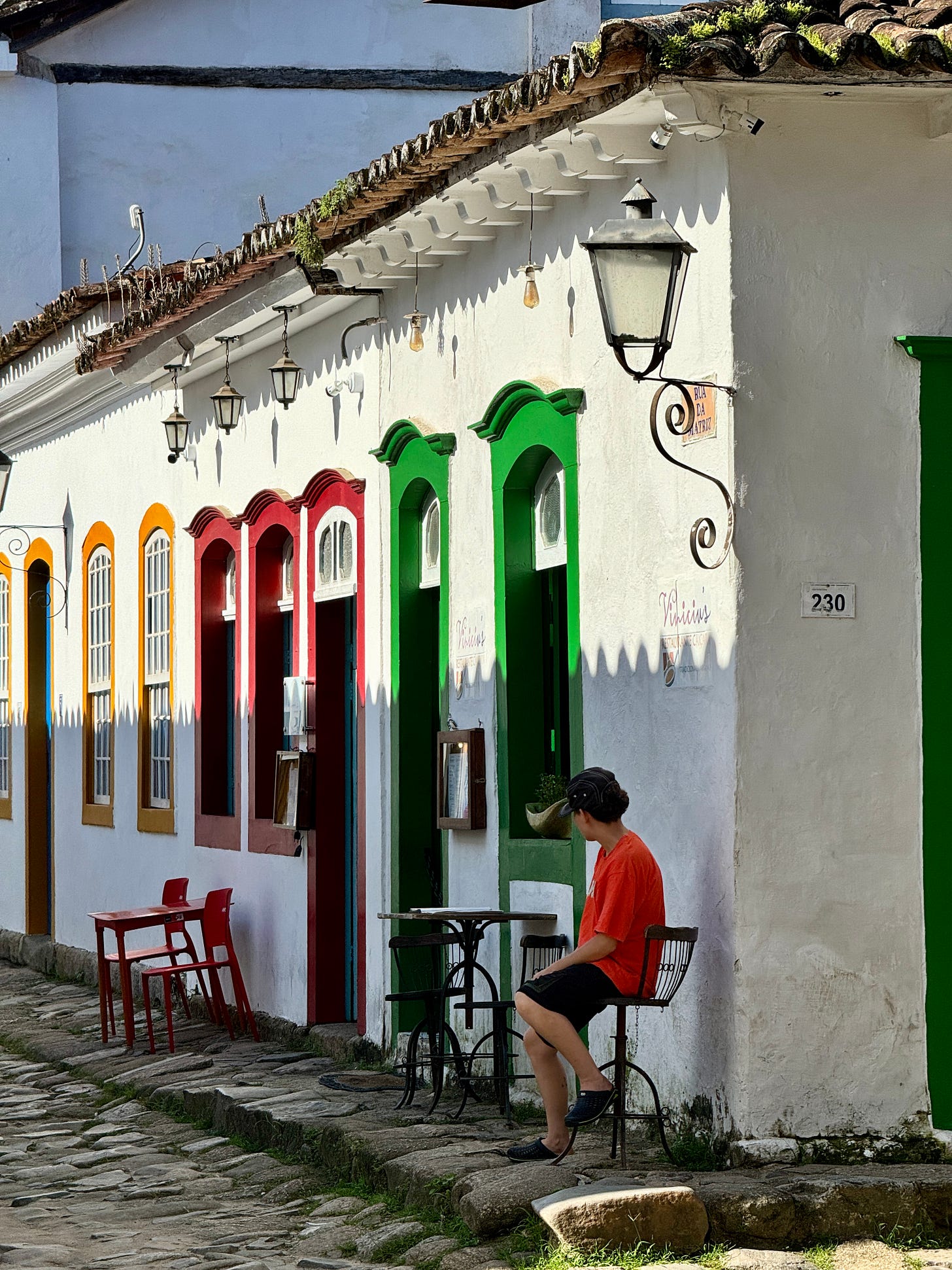 Chico sentado en banqueta en calle en Paraty. Calles adoquinadas, muros blancos y marcos de puerta de colores.