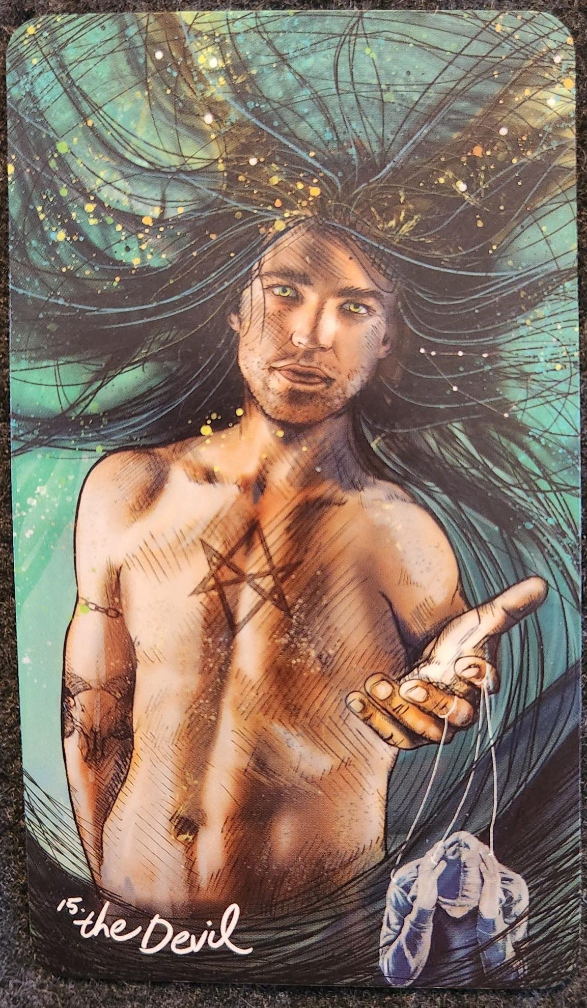 A tarot card labelled 15. The Devil. A shirtless, male-presenting person with long black hair, and a toned torso is reaching out one hand as though offering something to the reader. His face is neutral. Puppet strings are tied around two of his fingers. The strings lead to a figure wearing a hoodie with their head down, hands clasped around their eyes, as though distressed.