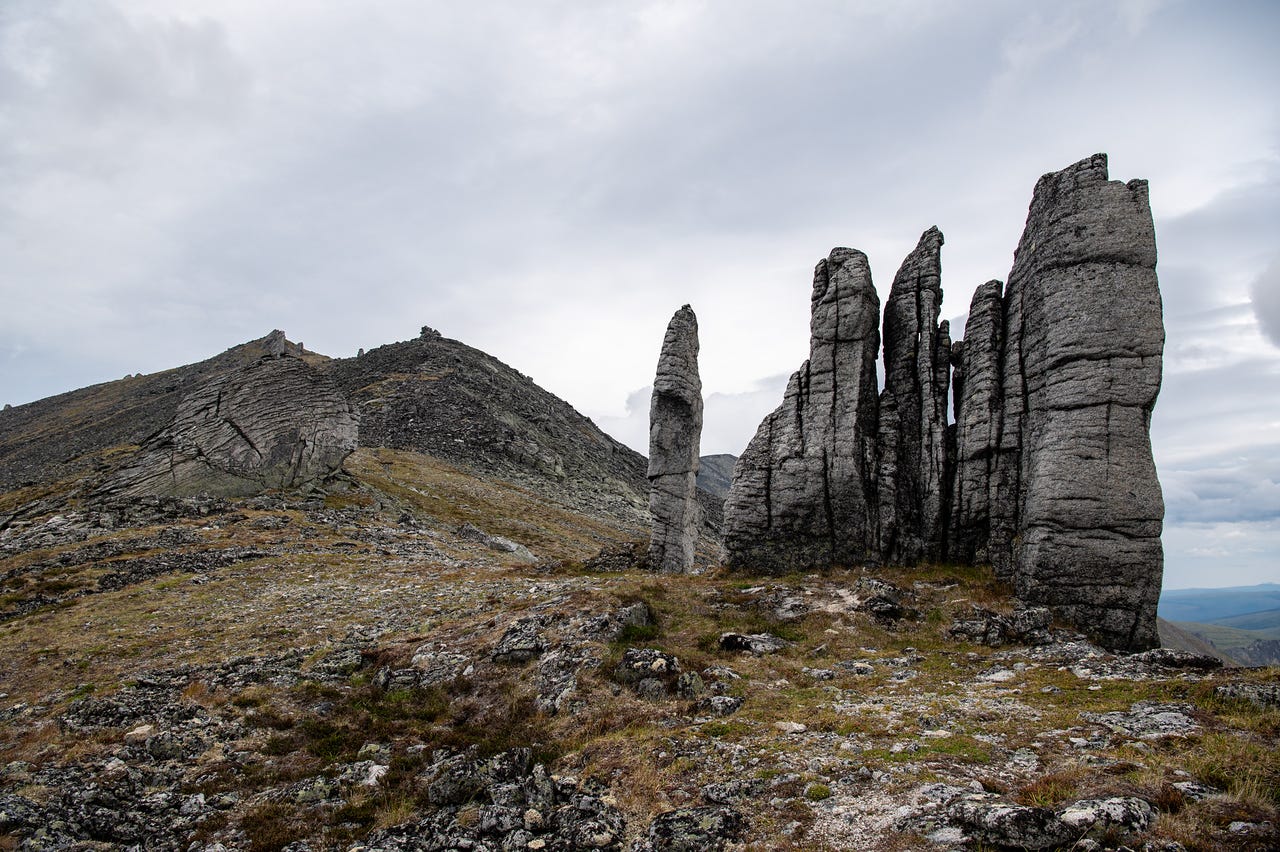 A cluster of tall, fluted granite columns stands on a rocky tundra ridge, weathered into vertical pillars streaked with dark lichen. A broad rubble-and-tundra slope climbs to a rounded summit at left under a pale grey sky.