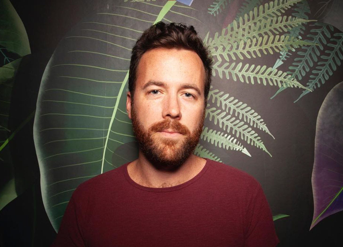 Rolling Stone culture writer Miles Klee wears a burgundy sweater and stands in front of wallpaper with ferns