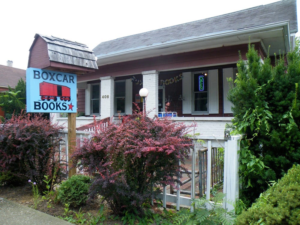 Shows the front of a house with porch. A sign out front says Boxcar Books with a blue background and a red icon of a train boxcar.