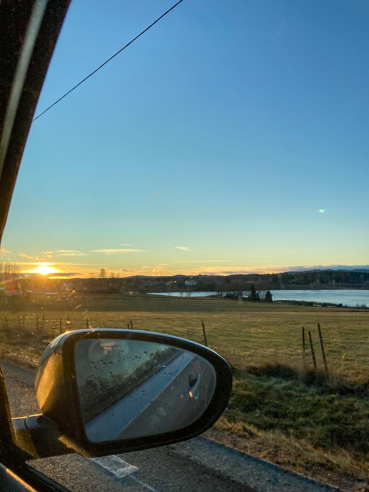 a person stands in the snow at the edge of a frozen lake, a person takes a photo out of a car window showing the wing mirror and the sun setting over a field