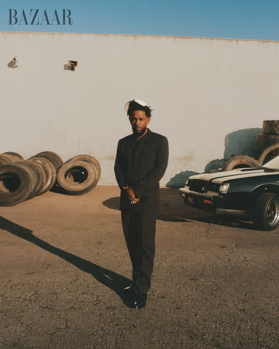 Kendrick Lamar wearing a black suit and standing in front of a large "BAZAAR" sign, a pile of old tires, and a vintage car.