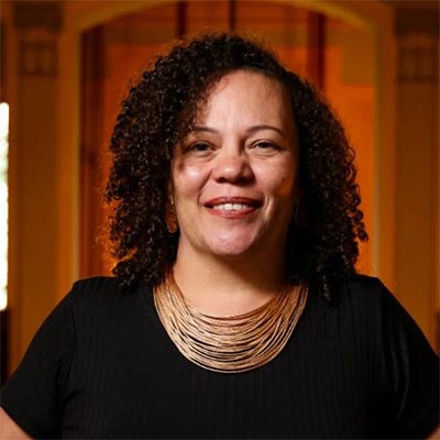 Mulher negra sorridente com cabelos cacheados na altura dos ombros. Ela veste uma camiseta preta e está em um ambiente interno com uma iluminação quente. Ela também utiiza brincos dourados e um colar douroado.