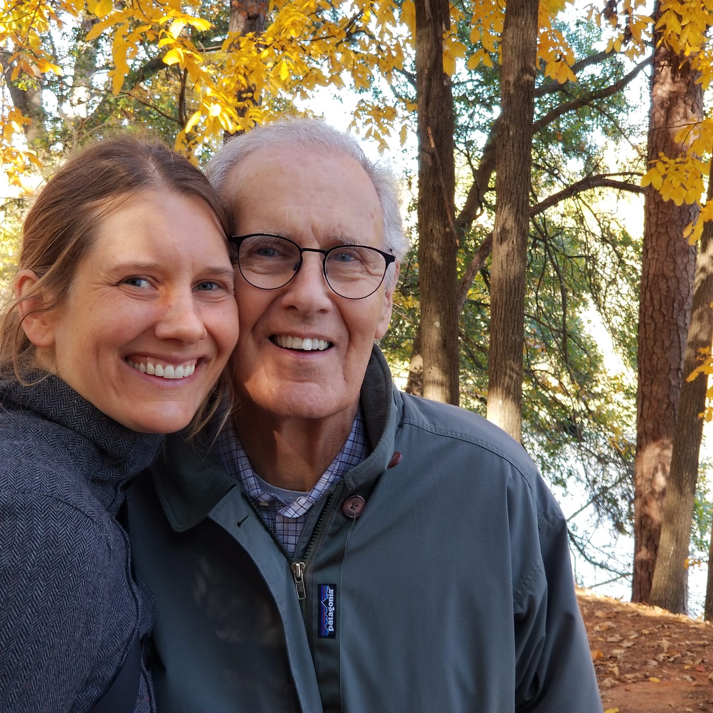 A younger woman and older man smile cheek-to-cheek in front of yellow autumn leaves