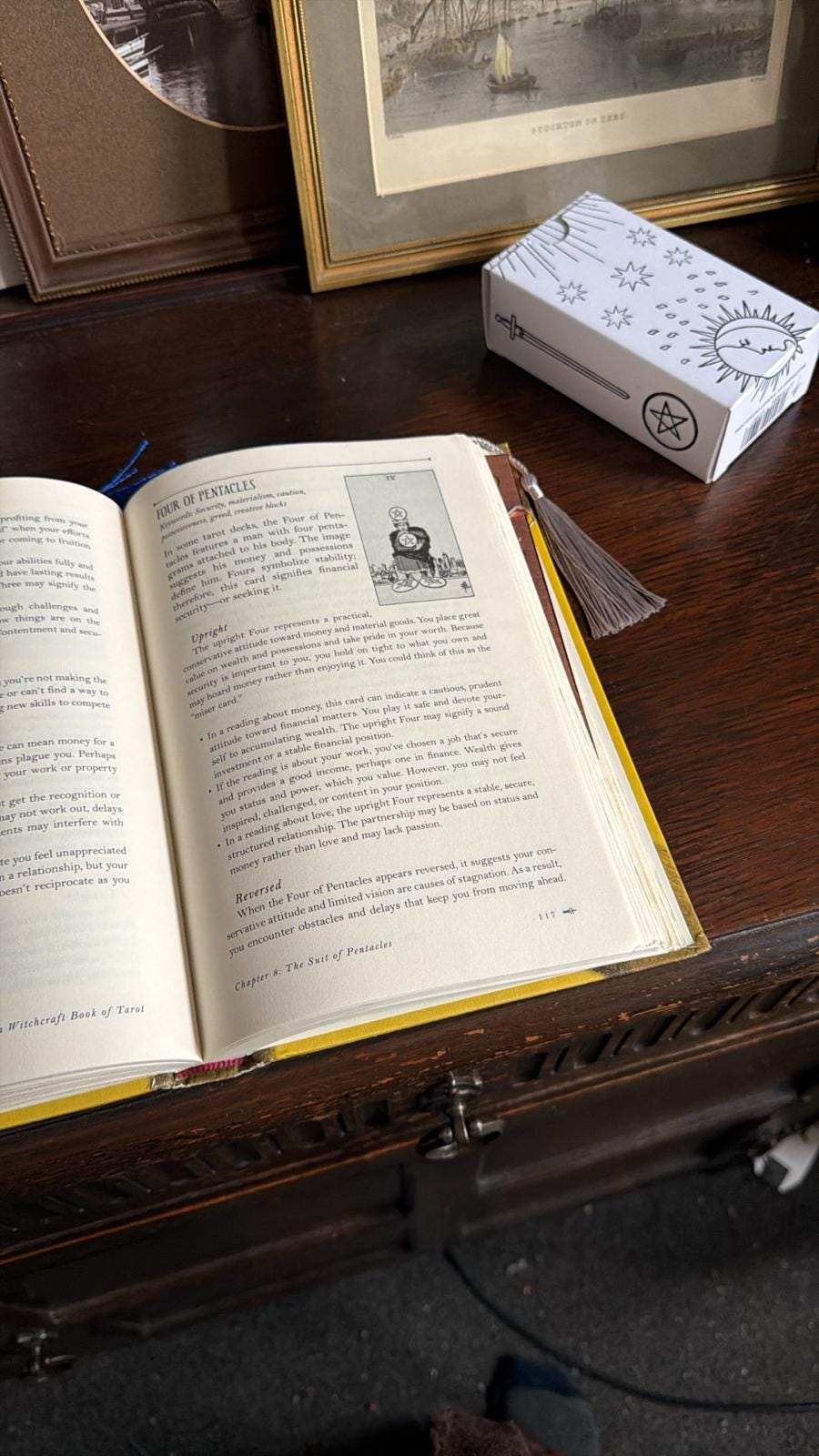 Open tarot reference book turned to the Four of Pentacles, shown beside a boxed tarot deck and tasselled bookmark on a wooden desk, illustrating the moment of synchronicity discussed in Lucy Rose’s Tarot DMs interview as she reflects on money, security, creative risk, and the realities of freelance writing after the success of The Lamb.