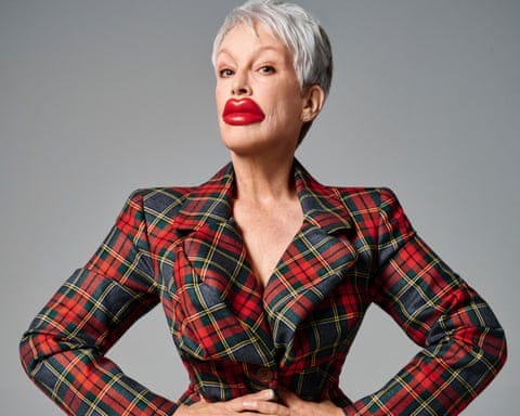 Portrait of Jamie Lee Curtis in a tartan jacket with her hands on her hips and sporting a huge pair of plastic red lips