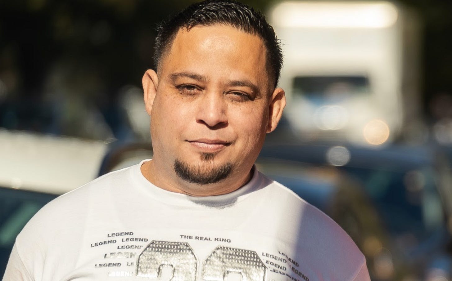 a brown-skinned man with short black hair and white t shirt smiles softly at the camera