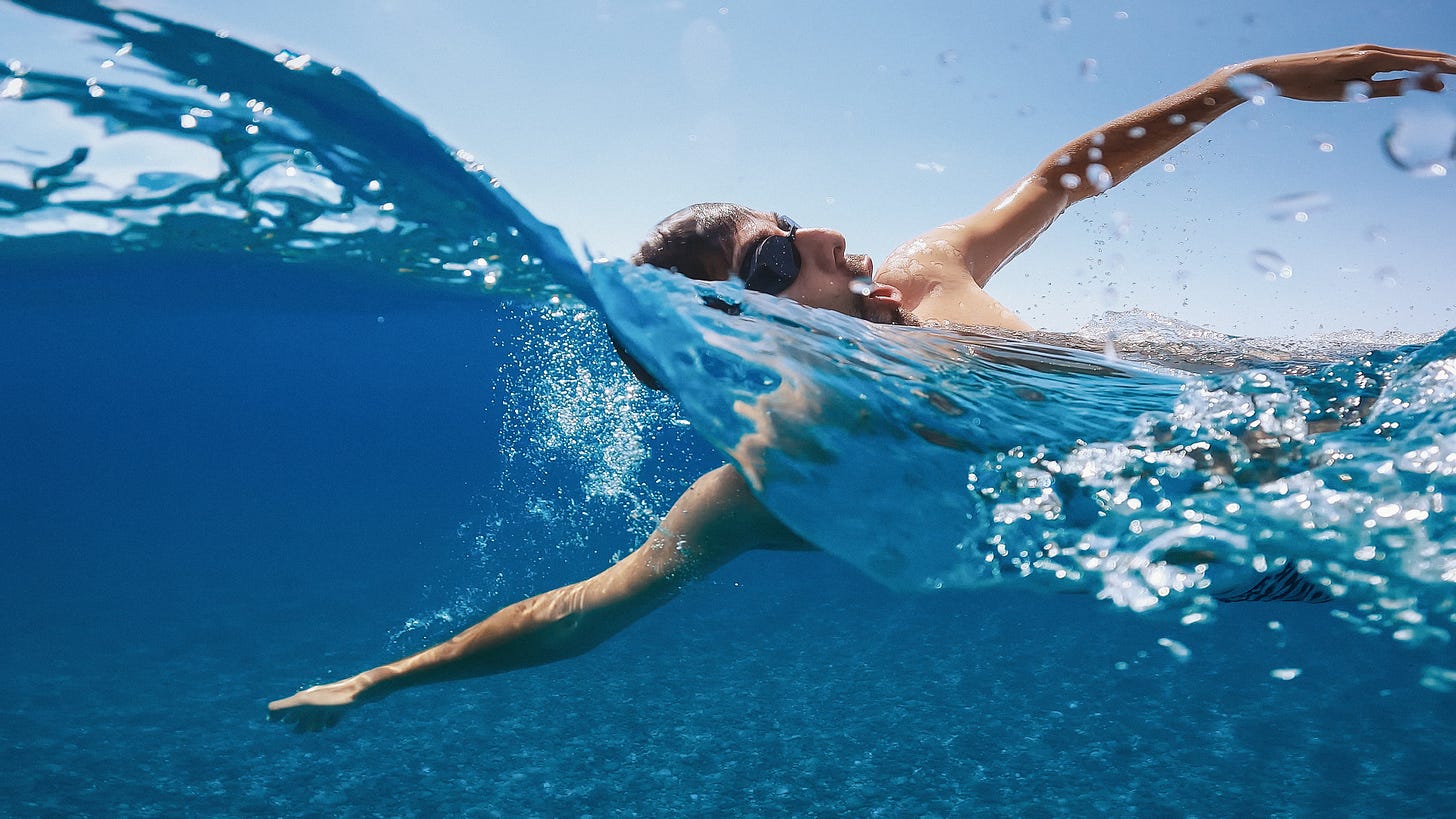 A person swims in a choppy ocean. They are midstroke, taking a breath. The water is a deep blue, and the sky a light blue. 