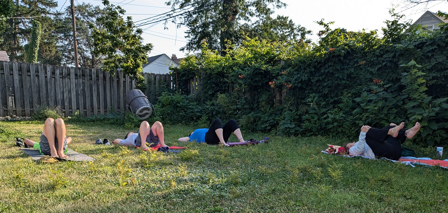 Four people lay on their backs on yoga mats in a back yard. There's a compost bin and a tall wooden fence behind the people, and along their left side, there's an overgrown border of trumpet vines covering another fence.