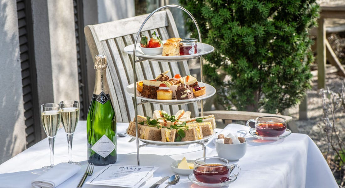 A tiered tray of afternoon tea on top of a white table with champagne and tea on it, next to a wooden chair outside