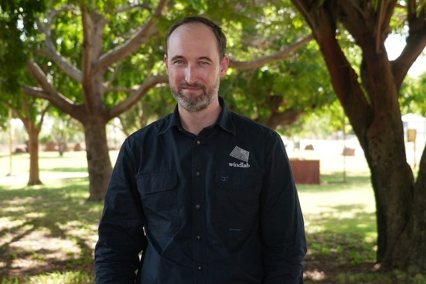 A man in a navy blue shirt stands in a park with trees in the back ground.