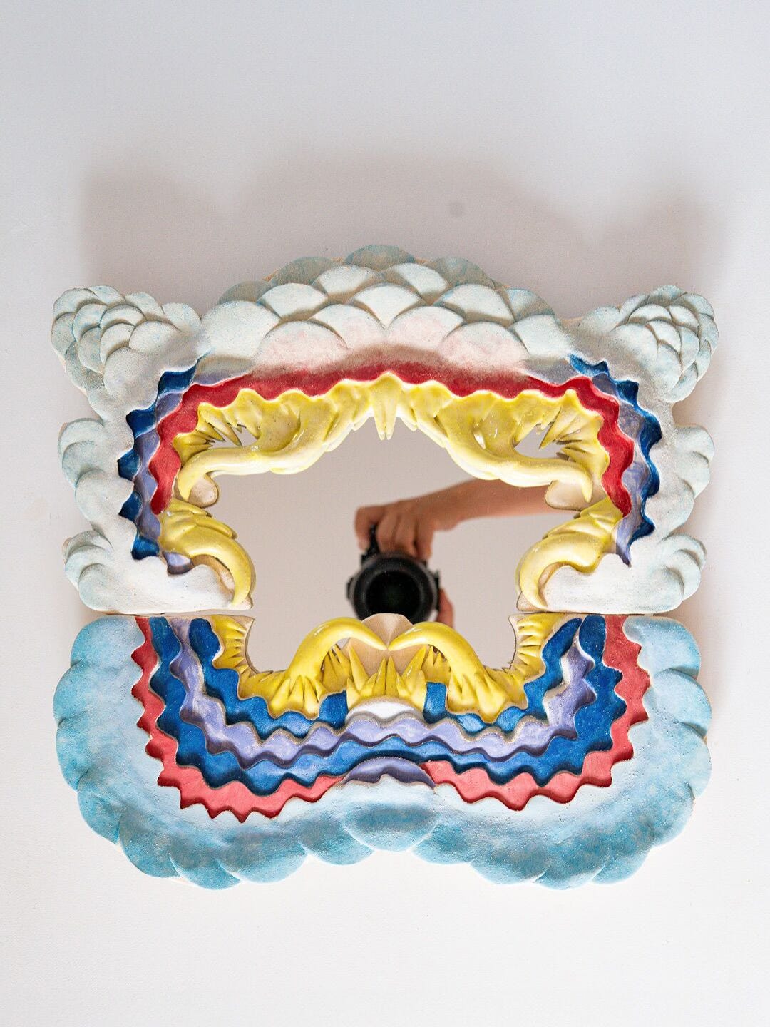 Eri Maeda’s 2025 artwork Monster Mirror, made of ceramic and mirror, merging reflective surface with monstrous ceramic forms to challenge beauty standards and self-perception. Colorful ceramic Monster Mirror by Eri Maeda with layered blue, red, yellow forms surrounding a central mirror that reflects the photographer holding a camera. Contemporary ceramic art, 2025.