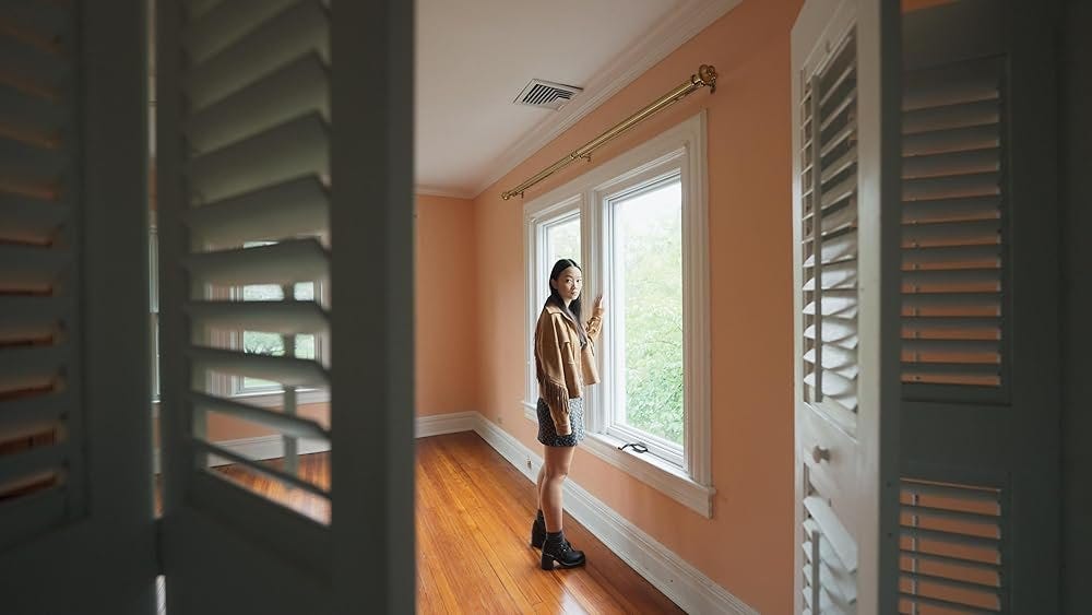 A view from inside a closet, looking out into an empty room with salmon-colored walls. A girl facing a large window turns to look into the camera.
